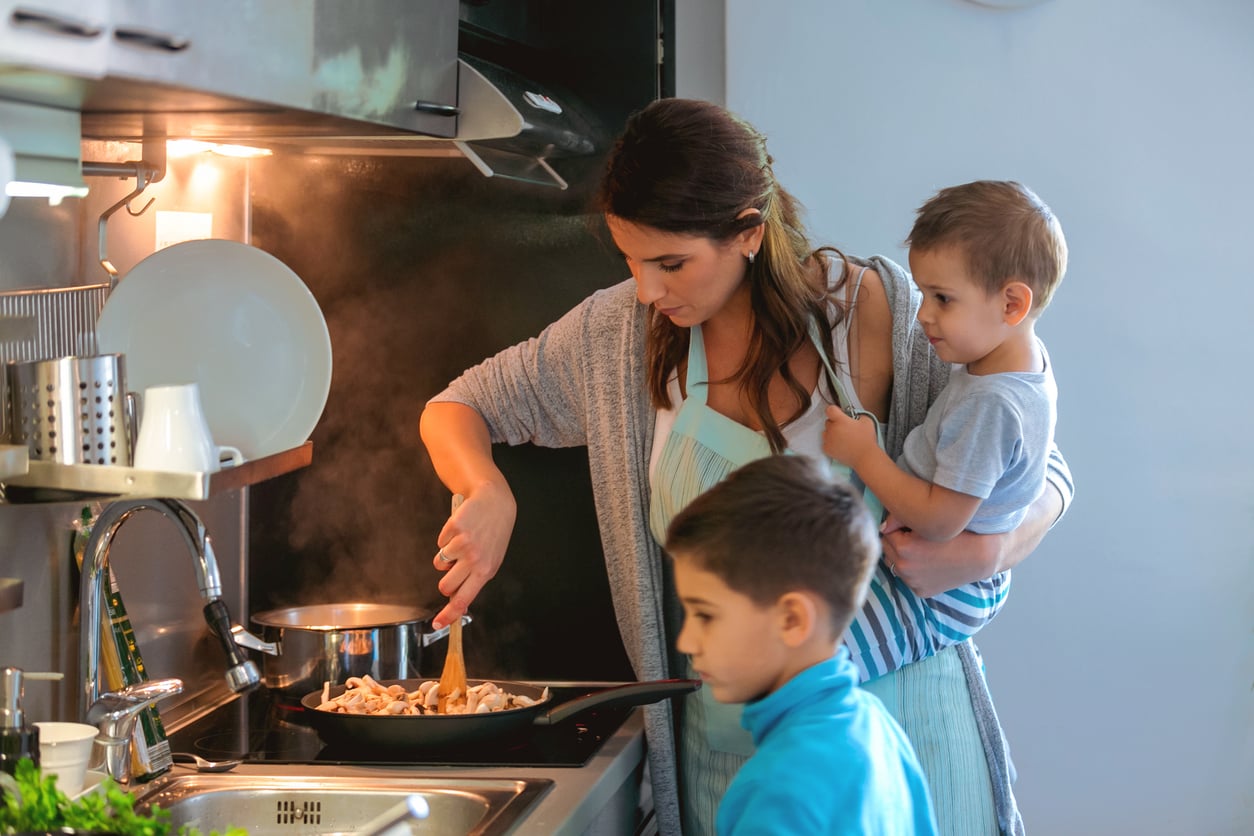 A woman cooks at a stove, stirring food in a pan while holding a young child on her hip. An older child stands beside her, watching. The kitchen is softly lit and steam rises from the pan.