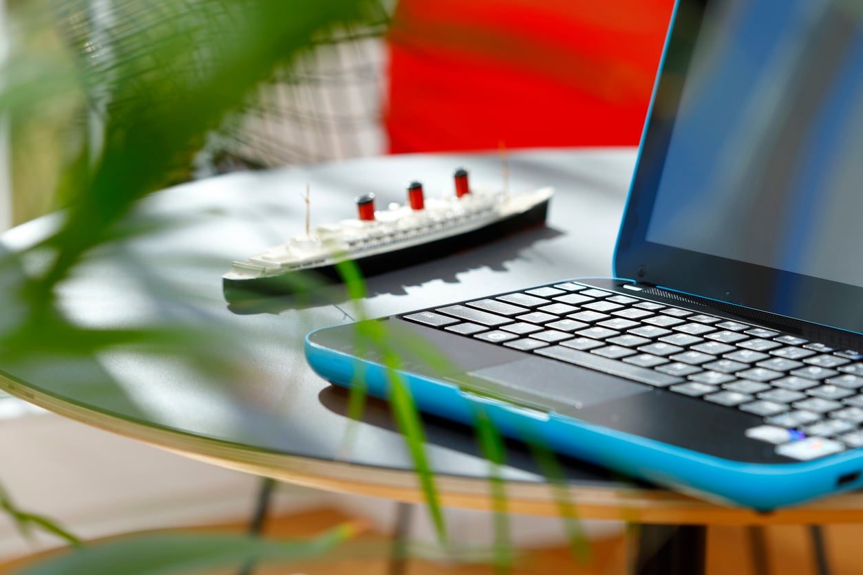 A close-up of a blue-edged laptop on a round table with a model ship in the background hints at travel hacks inspiration. Green leaves partially obscure the view, adding to the bright, casual setting.