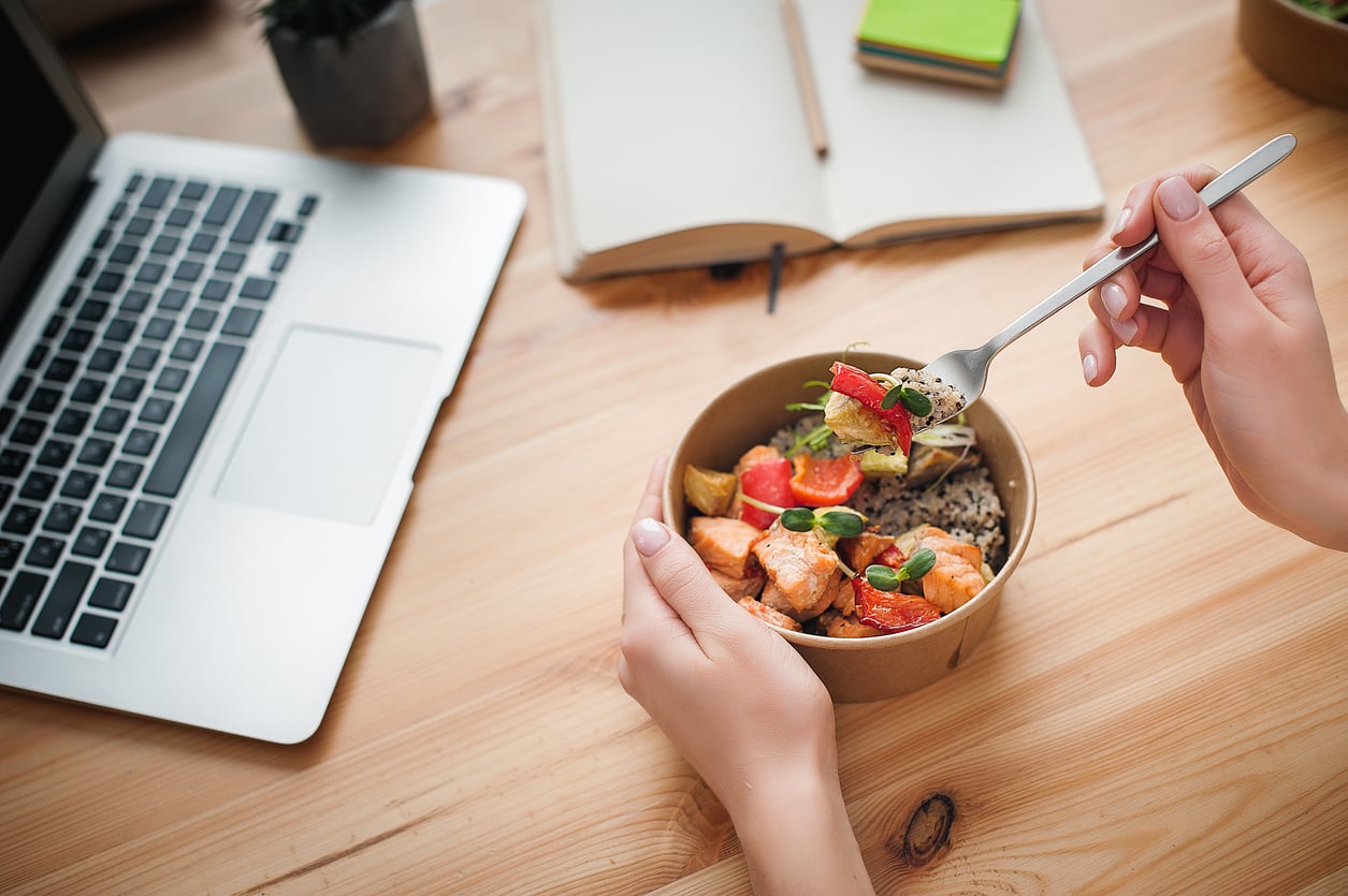 A person eating a healthy meal with vegetables and chicken from a bowl at a desk, next to a laptop, open notebook, and stacked notepads. Spending habits in 2026.
