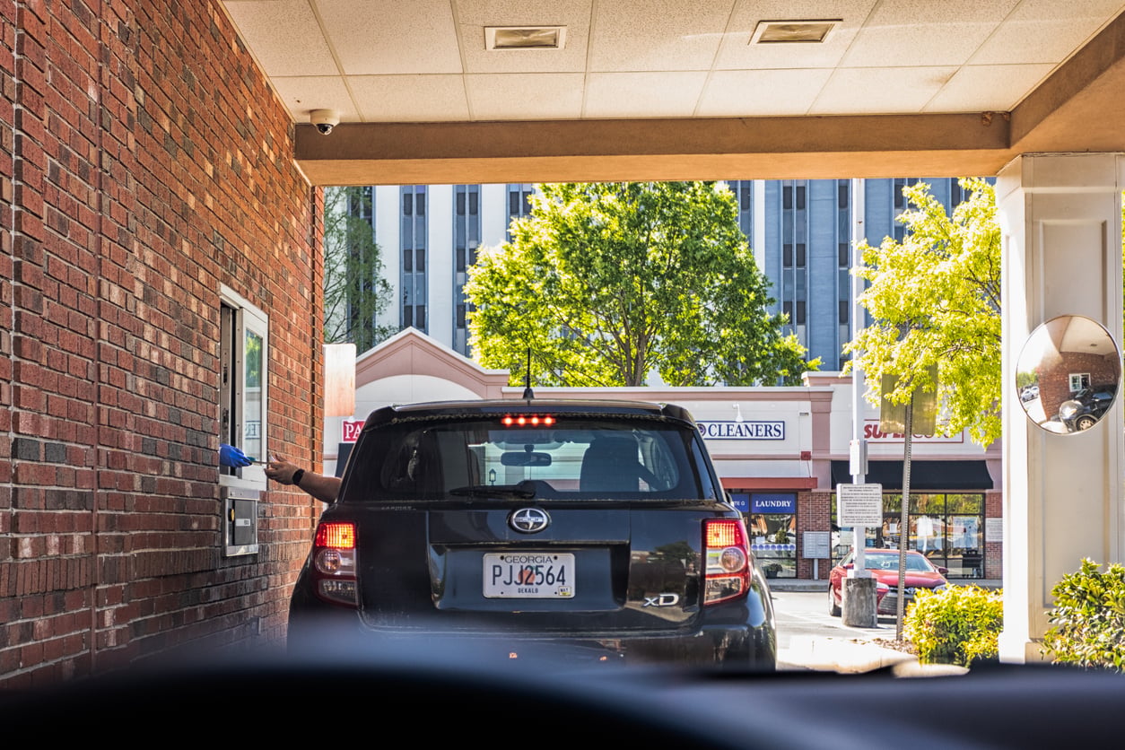 A black SUV with a Florida license plate waits at a drive-thru window, where a gloved employee reaches out. A brick wall lines the left, and trees and buildings are visible outside in bright daylight.