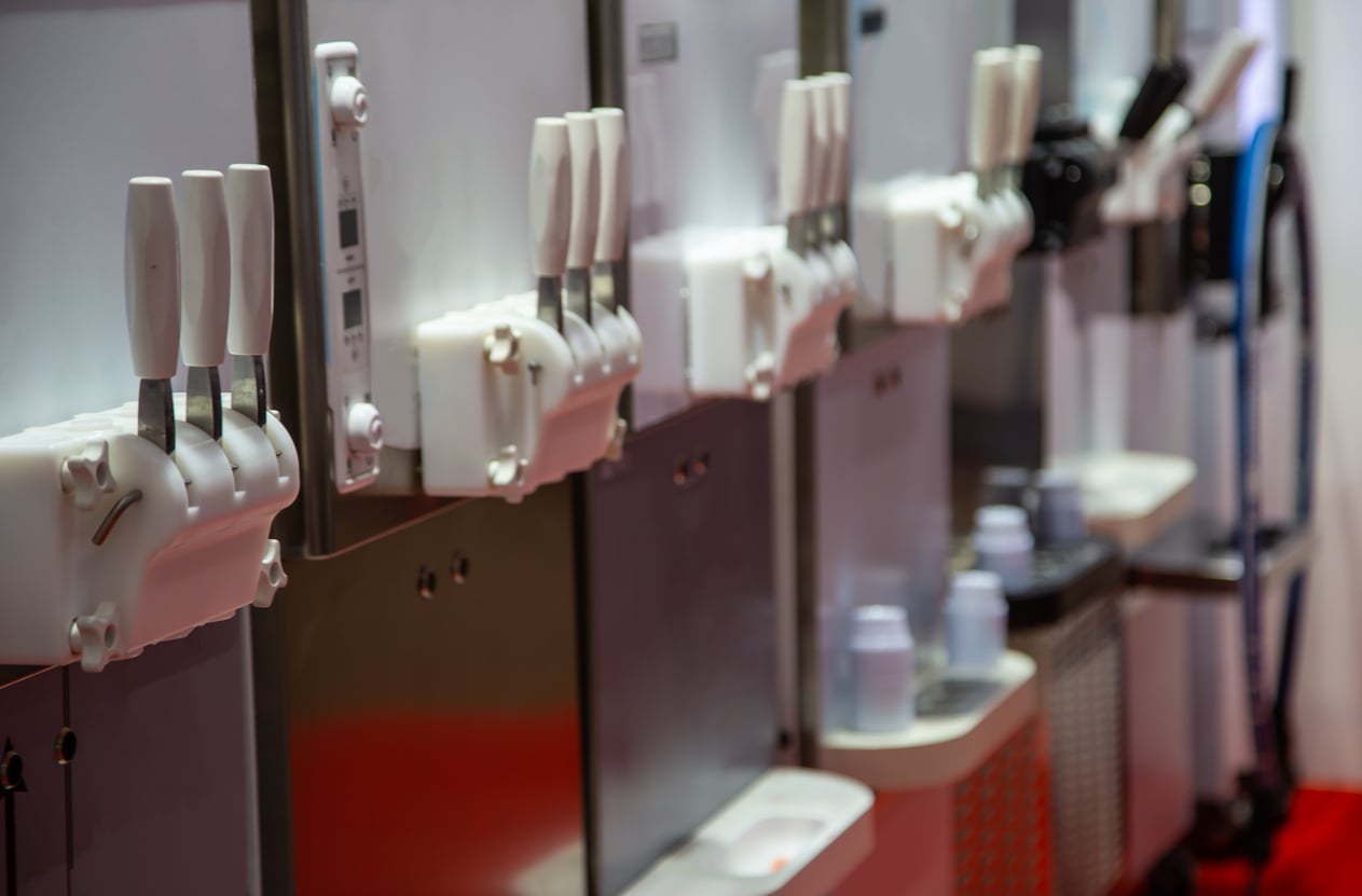 Close-up of several soft serve ice cream machines with white levers and empty cups placed on a shelf below the dispensers. The background is blurred, focusing on the machine handles.