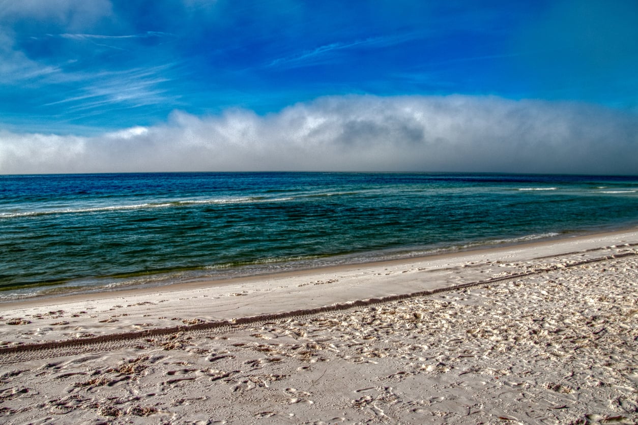 A sandy beach with footprints leads to clear blue-green ocean water under a bright blue sky with light clouds on the horizon.