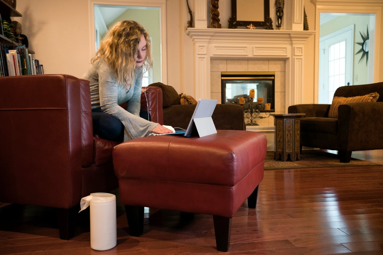 A woman sits on a red chair in a living room, using a tablet on a matching ottoman. Disinfectant wipes are visible on the floor, and the room has a fireplace, wood floors, and cozy décor.