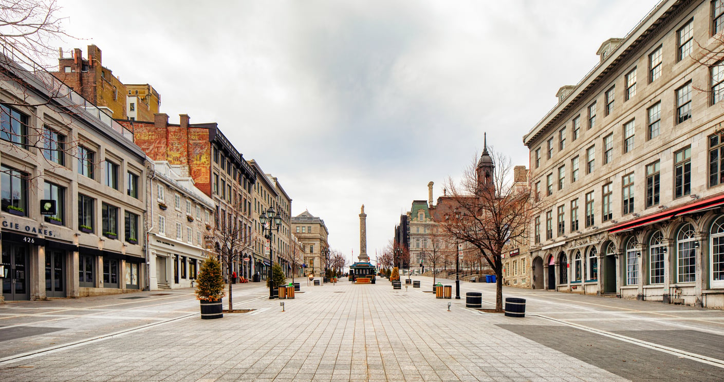 A wide, empty cobblestone street flanked by historic buildings on both sides, with a monument and statue visible in the distance beneath a cloudy sky.
