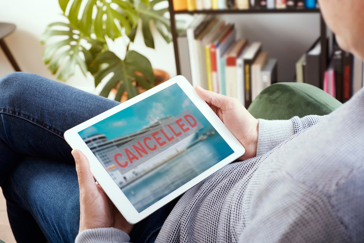 A person holds a tablet displaying an image of a cruise ship with "CANCELLED" in bold red letters. In the background, bookshelves and a plant suggest they might be staying at an Airbnb instead.