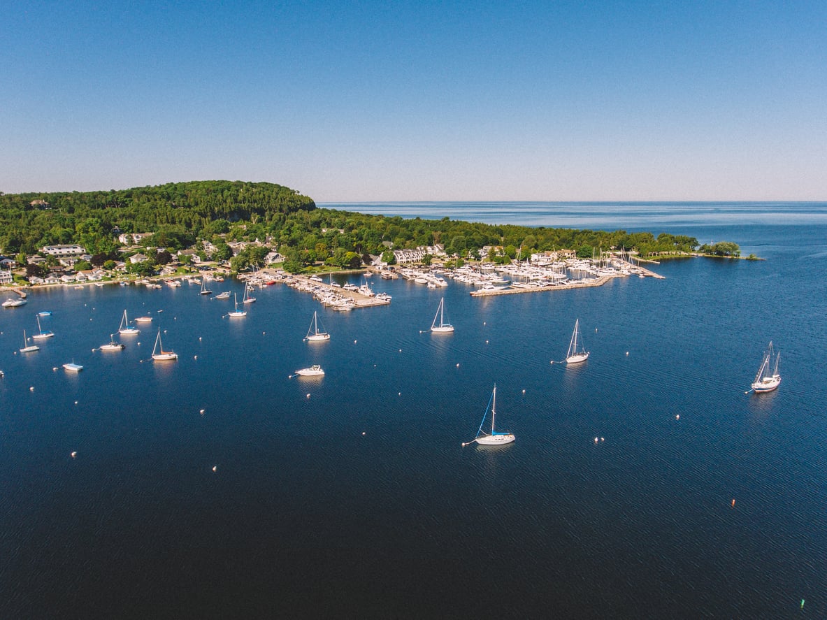 Aerial view of a calm harbor with several sailboats anchored in blue water, a tree-covered shoreline, marina, and buildings along the coast under a clear blue sky.