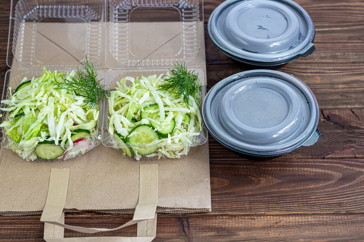 Two plastic containers with fresh cucumber and cabbage salad, garnished with dill, sit on a brown paper bag next to two closed, round plastic containers—an appetizing scene often found among overpriced products on a wooden table.