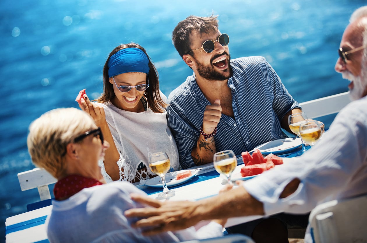 Four people sit at an outdoor table by the ocean, laughing and enjoying drinks and watermelon under the sun. Wearing sunglasses and light clothing, they embody the cheerful, relaxed vibe of a Best Cruise Line 2026 getaway.
