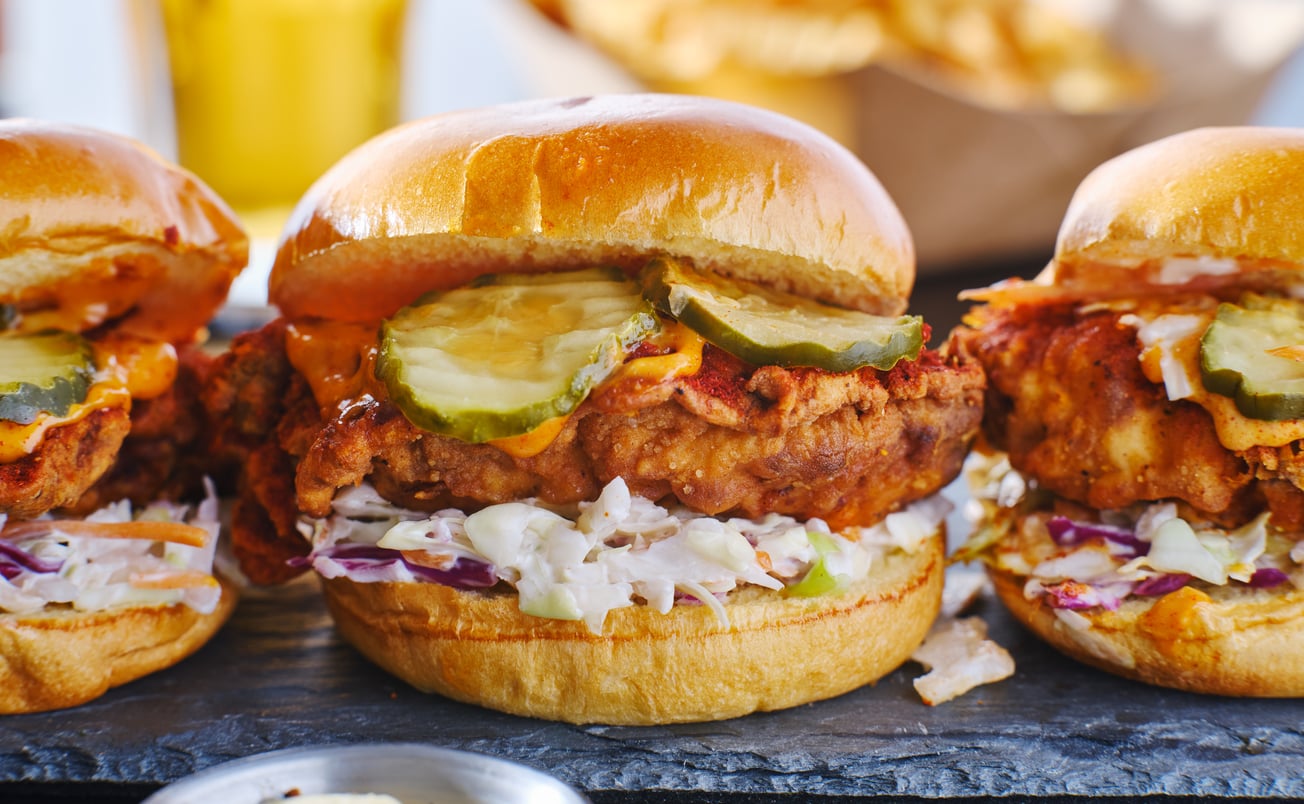 Three crispy fried chicken sandwiches on brioche buns, topped with pickles, coleslaw, and sauce, are arranged on a slate board with fries and a drink in the blurred background.