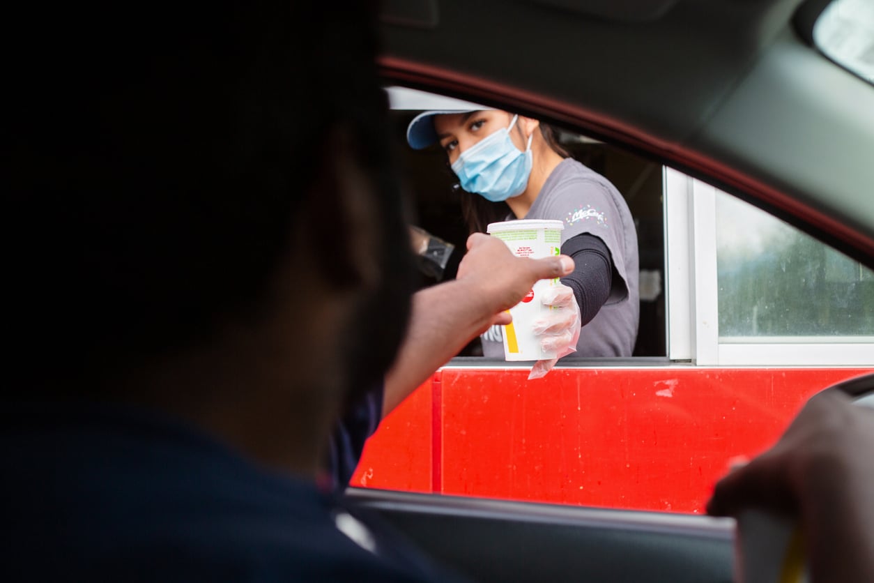 A fast food worker wearing a mask hands a drink to a customer through a drive-thru window. The customer is reaching out from inside their car to take the cup.