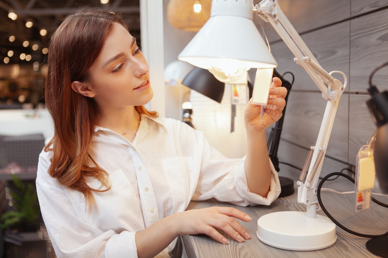 A woman in a white shirt examines a price tag on a white desk lamp in a store, surrounded by other lamps on display. How tariffs affect consumers. 