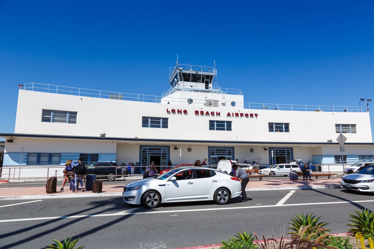 Travelers stand outside the main terminal of Long Beach Airport, one of the most convenient airports, with parked cars, luggage, and a clear blue sky overhead.