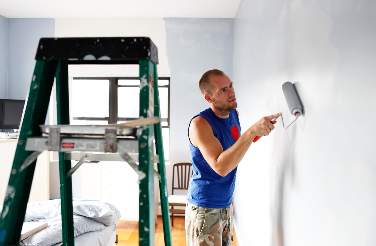 Demonstrating his DIY skills, a man in a blue sleeveless shirt and camouflage shorts paints a light blue wall with a roller in a brightly lit room; a ladder and chair are nearby.