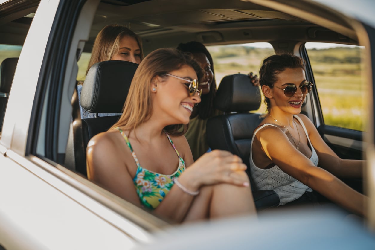 Four friends sit inside a car, smiling and laughing together. Two women are in the front seats, both wearing sunglasses, with sunlit scenery visible through the windows.