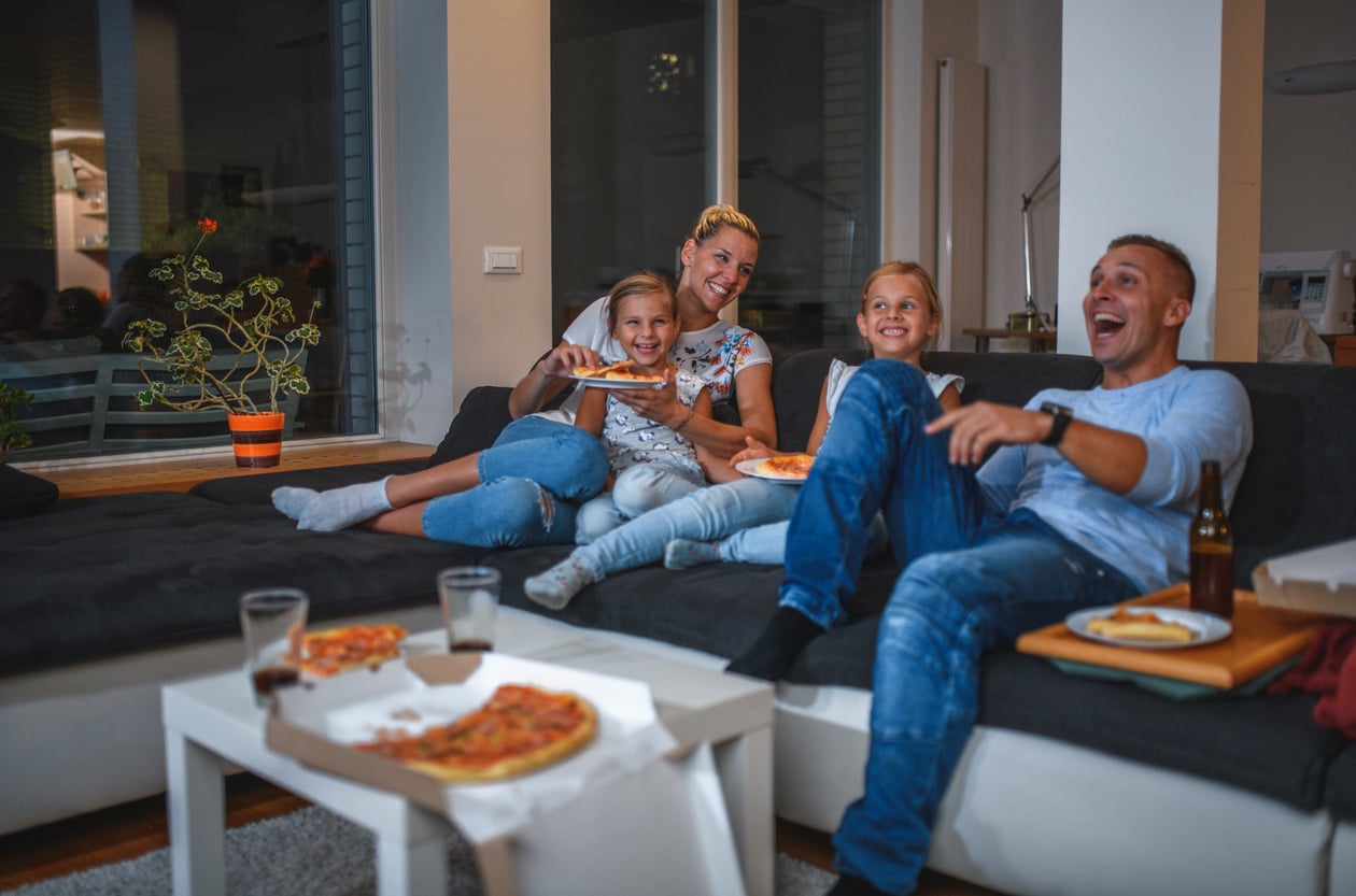 A family of four sits on a couch in a cozy living room, smiling and eating pizza while watching TV. Surrounded by pizza boxes and drinks, they enjoy a relaxing evening together—almost like enjoying 1991 prices for a night in.