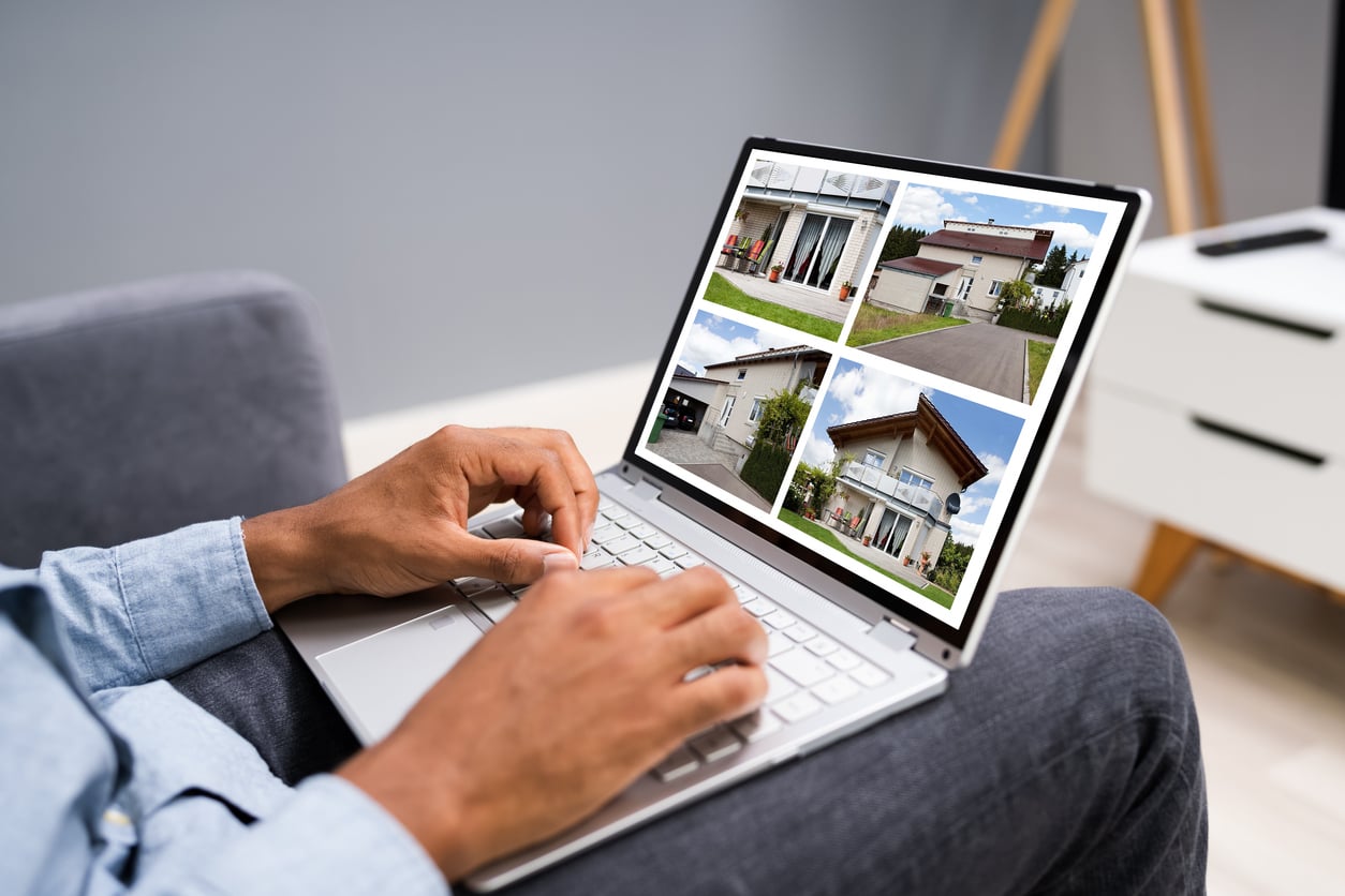 A person using a laptop, browsing real estate listings with four different houses displayed on the screen. The simple indoor background hints they're staying at an Airbnb, with only their hands and part of their lap visible.