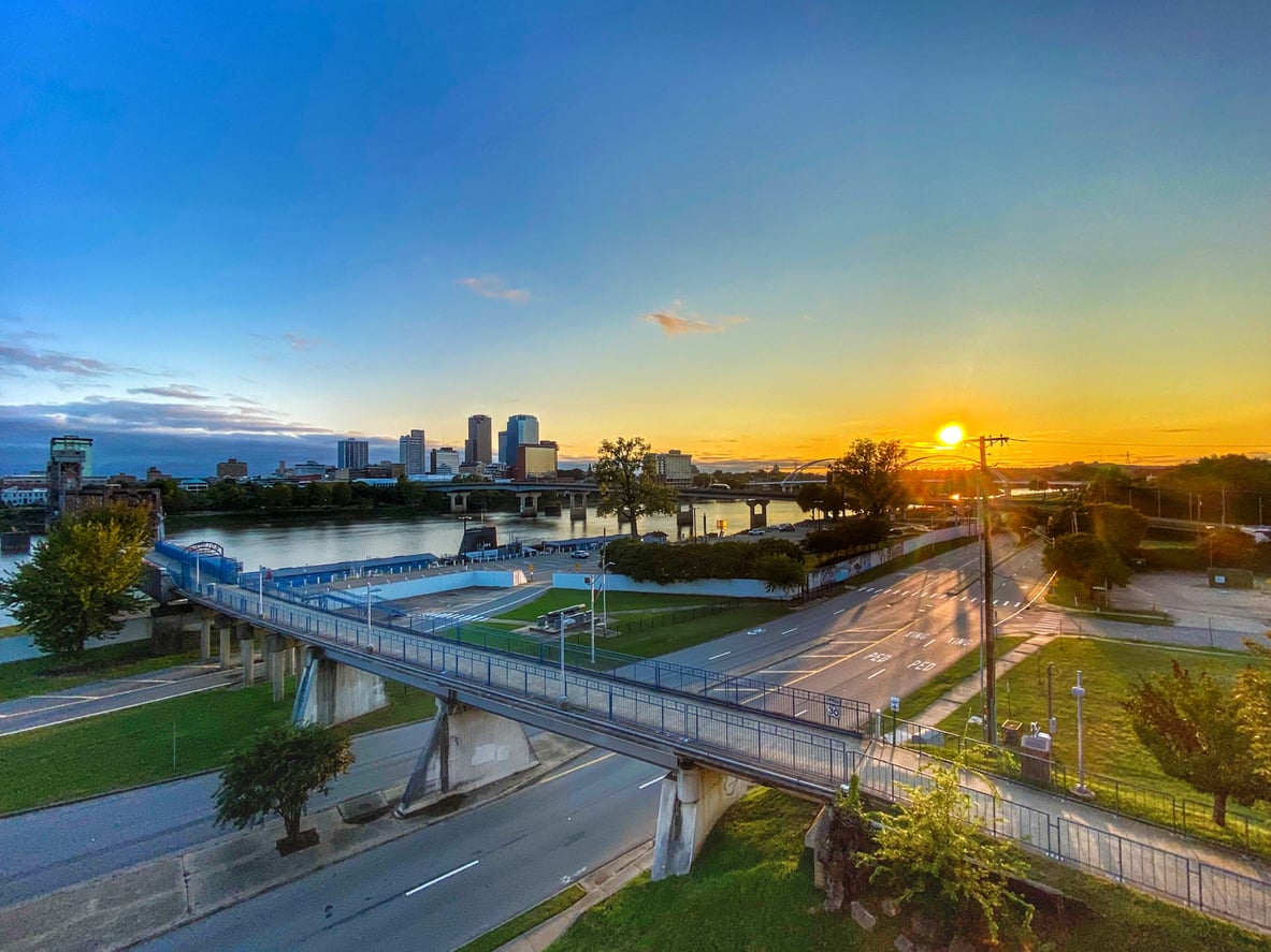 A vibrant sunset over a city skyline with a river, bridge, and park in the foreground. The sky is mostly clear with a few clouds and the sun is low near the horizon, casting a warm golden light.