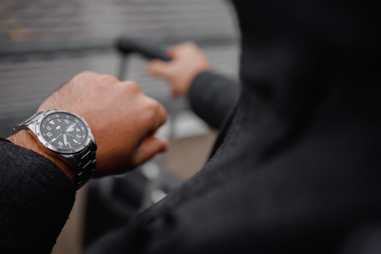 A person wearing a black hoodie looks at a silver wristwatch—one of those classic products men still buy—while holding the handle of a suitcase or stroller, with the background blurred.