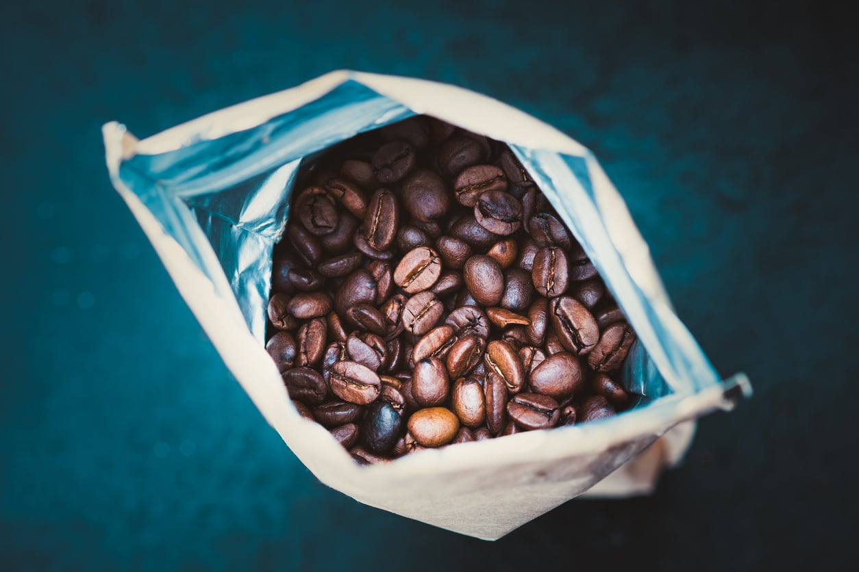 A close-up, top-down view of an open paper bag filled with dark roasted coffee beans, with the shiny inner lining of the bag visible against a dark, blurred background.