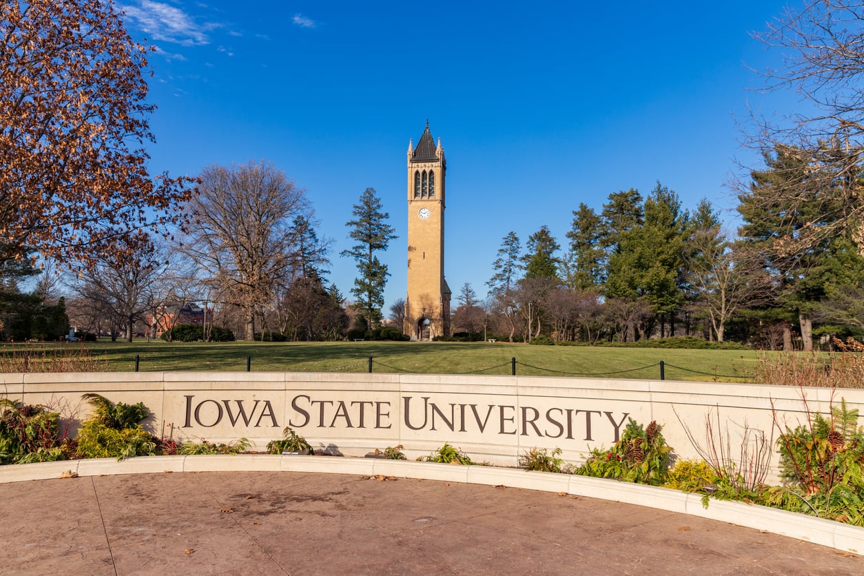 A large sign reads "Iowa State University" in front of a grassy lawn and the Campanile clock tower, surrounded by trees under a clear blue sky—showcasing one of the most affordable college towns.