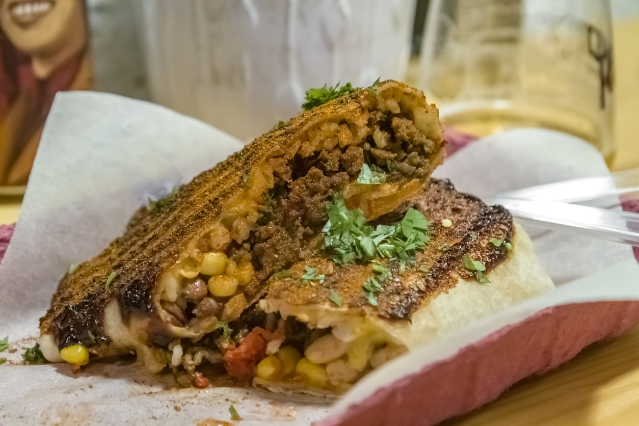 A close-up of a grilled taco or quesadilla cut in half, revealing a filling of ground meat or Ground Beef Alternatives, corn, beans, melted cheese, and chopped cilantro, served on parchment paper with a drink in the background.