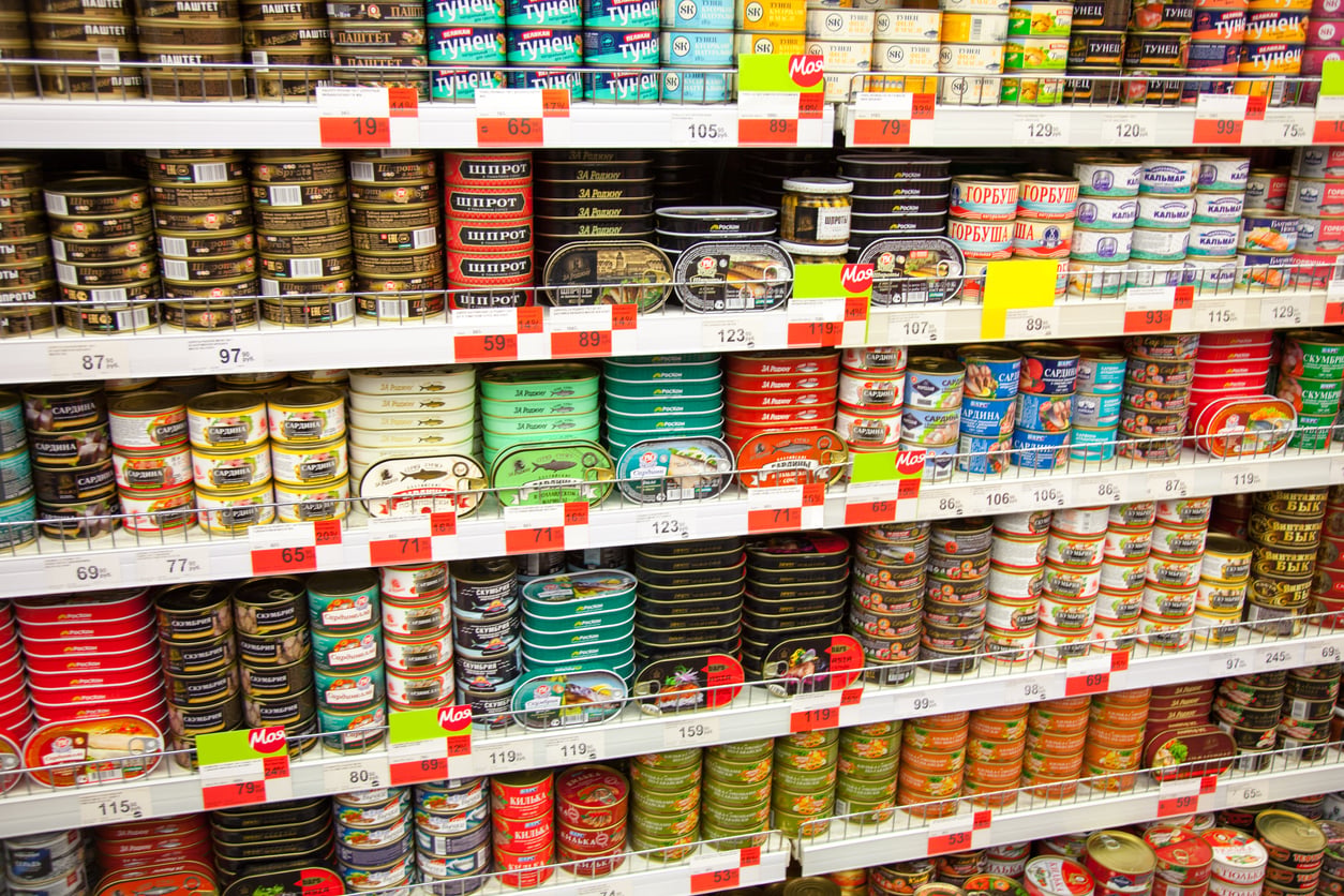Shelves in a grocery store stocked with various brands and types of canned fish in colorful packaging, each with price tags displayed below the products, alongside a selection of ground beef alternatives.