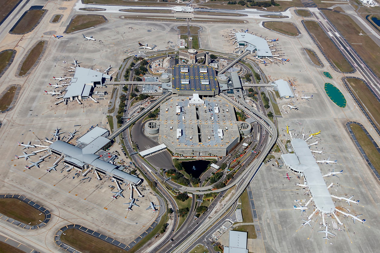 Aerial view of a large, convenient airport terminal with multiple concourses. Several airplanes are parked at gates, taxiways and runways surround the terminal, and service roads connect different parts of the airport complex.