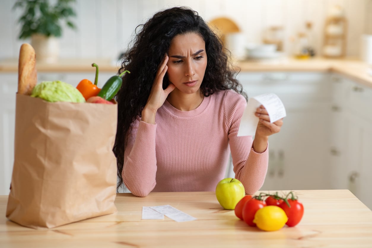 A woman with long curly hair looks worried while reading a receipt at a kitchen table, with groceries including bread, vegetables, and fruit beside her.