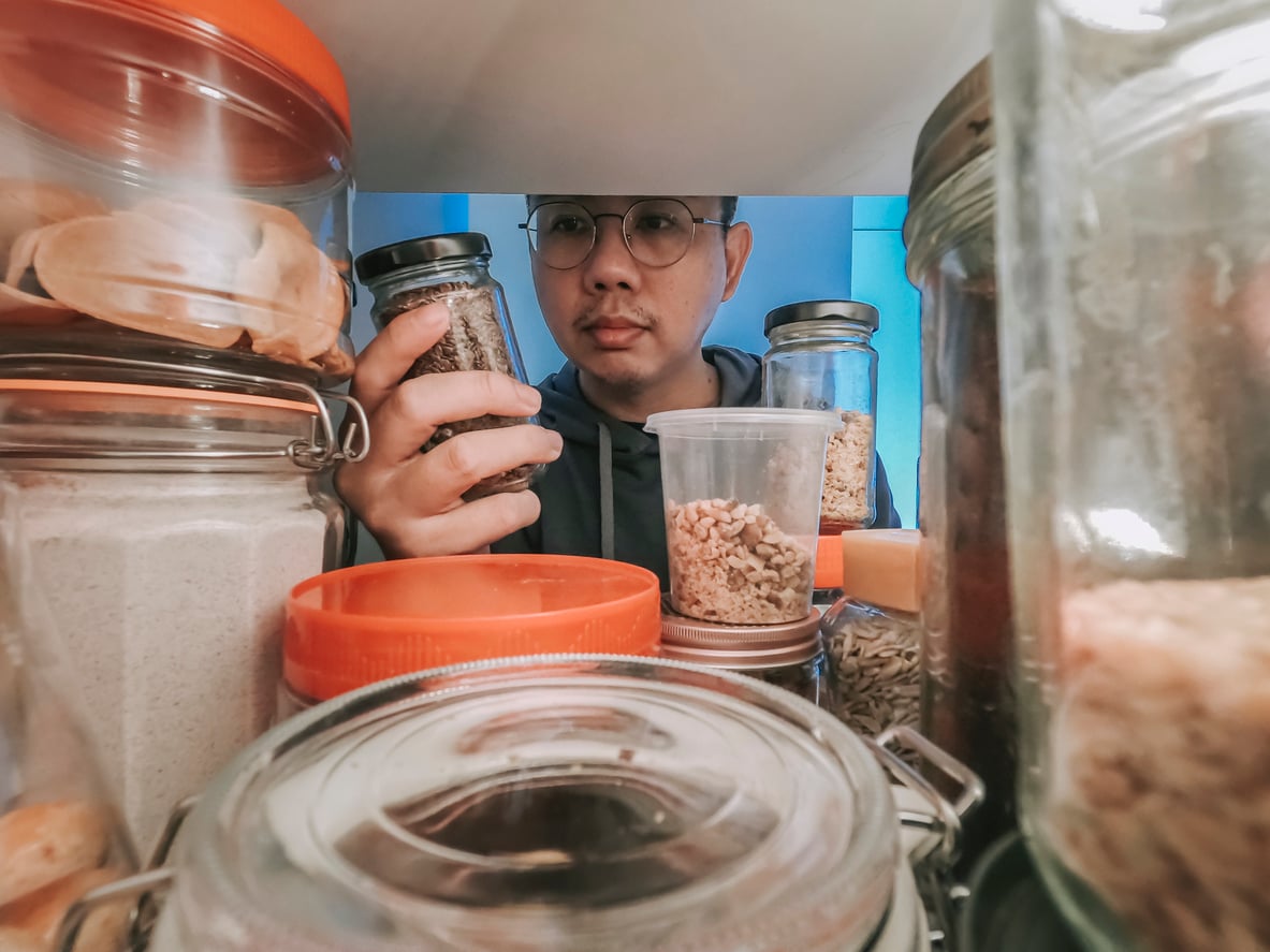 A person wearing glasses looks at a jar of food while standing in front of a shelf filled with various jars and containers, seen from inside the pantry or cupboard.