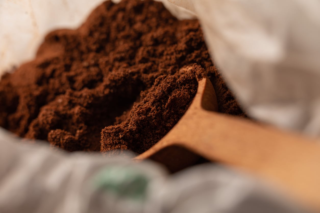 Close-up of a wooden spoon scooping finely ground coffee from a white bag. The coffee grounds are dark brown and textured, filling most of the frame.