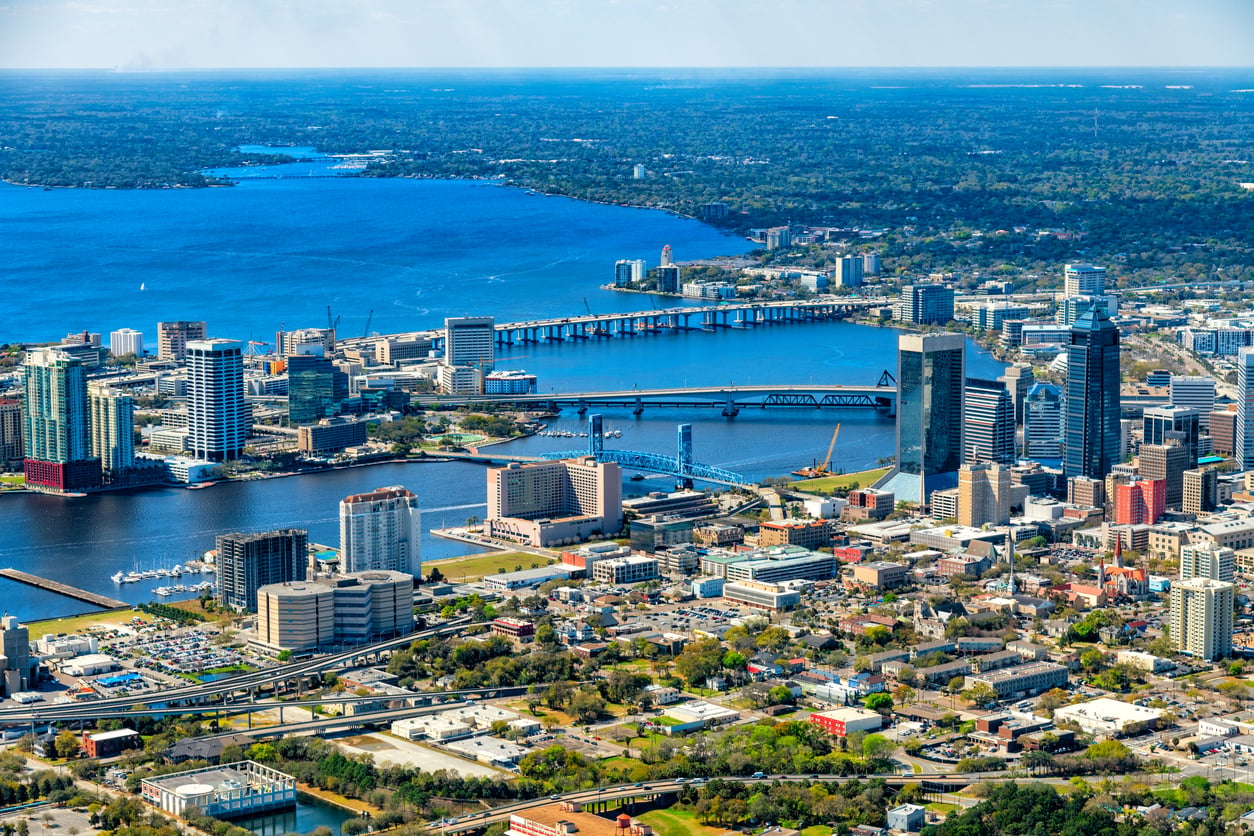 Aerial view of downtown Jacksonville, Florida, showing tall buildings, bridges, and the St. Johns River with sprawling urban and green areas in the background.