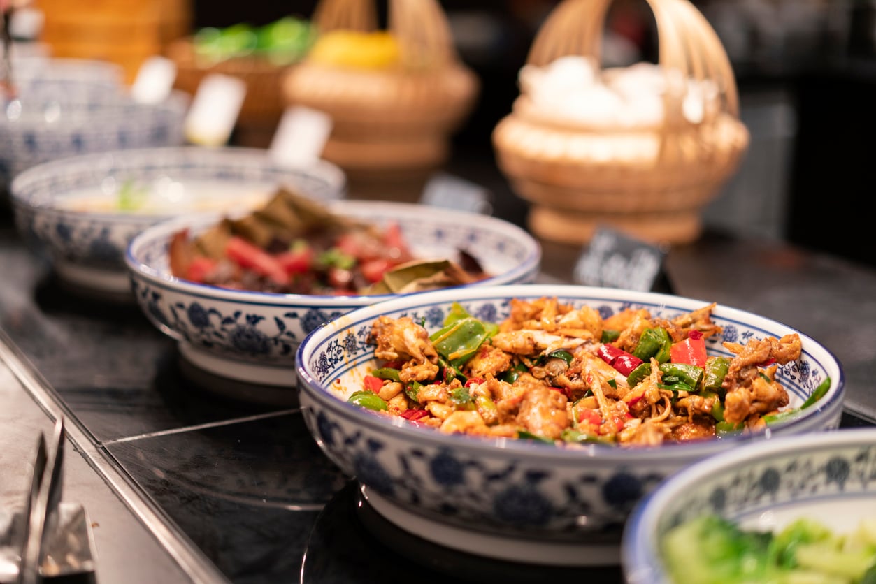 A close-up of a buffet with large decorative bowls filled with colorful stir-fried dishes, featuring vegetables and meat. Wicker baskets and more food are visible in the background, slightly out of focus.