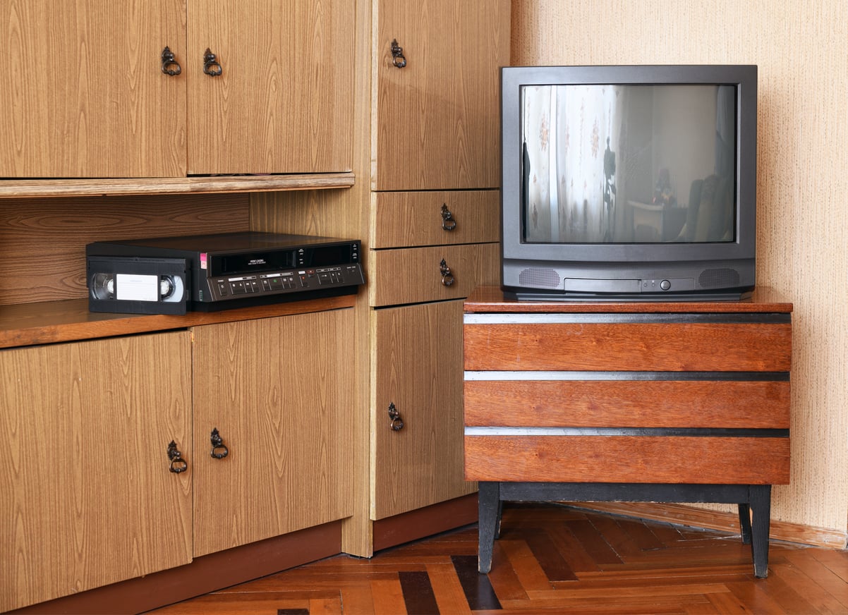 A vintage CRT television sits on a wooden stand next to a cabinet with a VHS player and a VHS tape inside, in a room with parquet flooring and beige walls. Cost of living in the 1980s