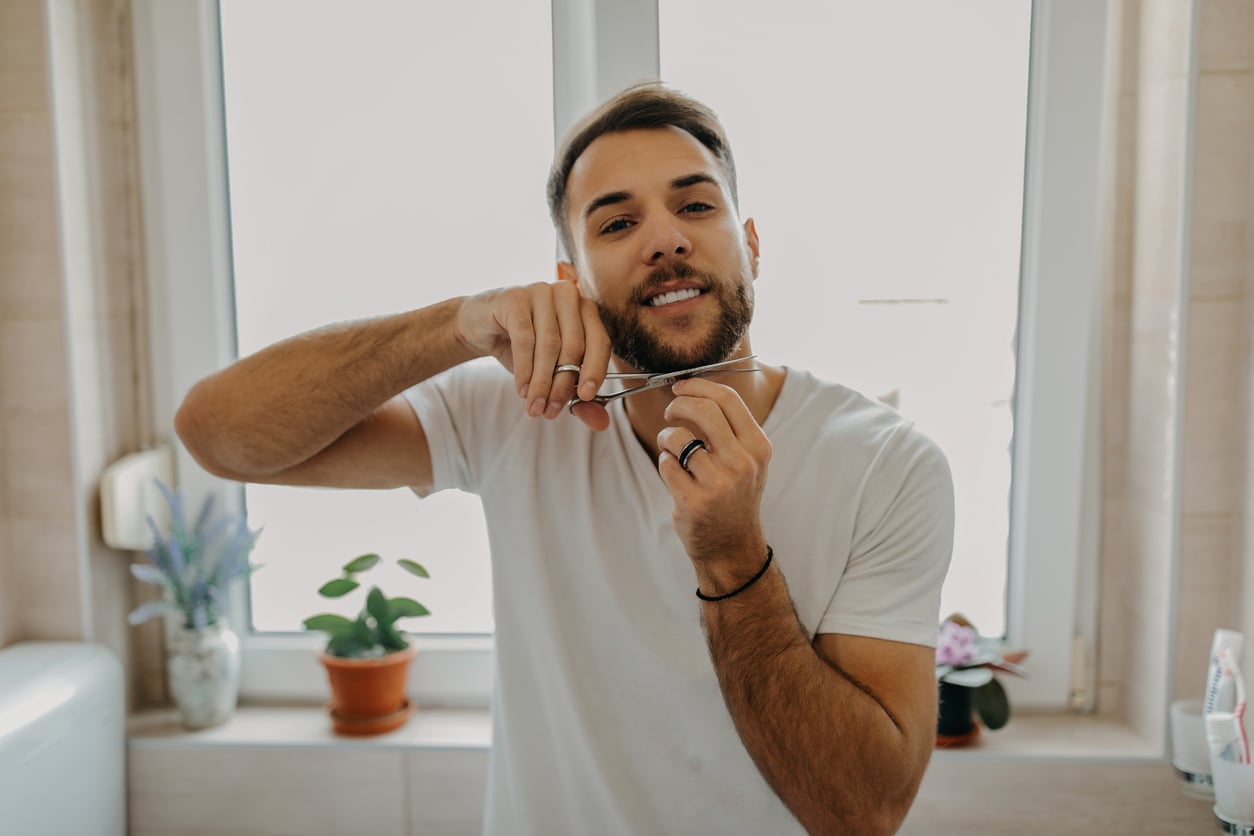 A man in a white t-shirt showcases his DIY skills as he trims his beard with scissors, smiling in a bright bathroom with potted plants on the windowsill behind him.