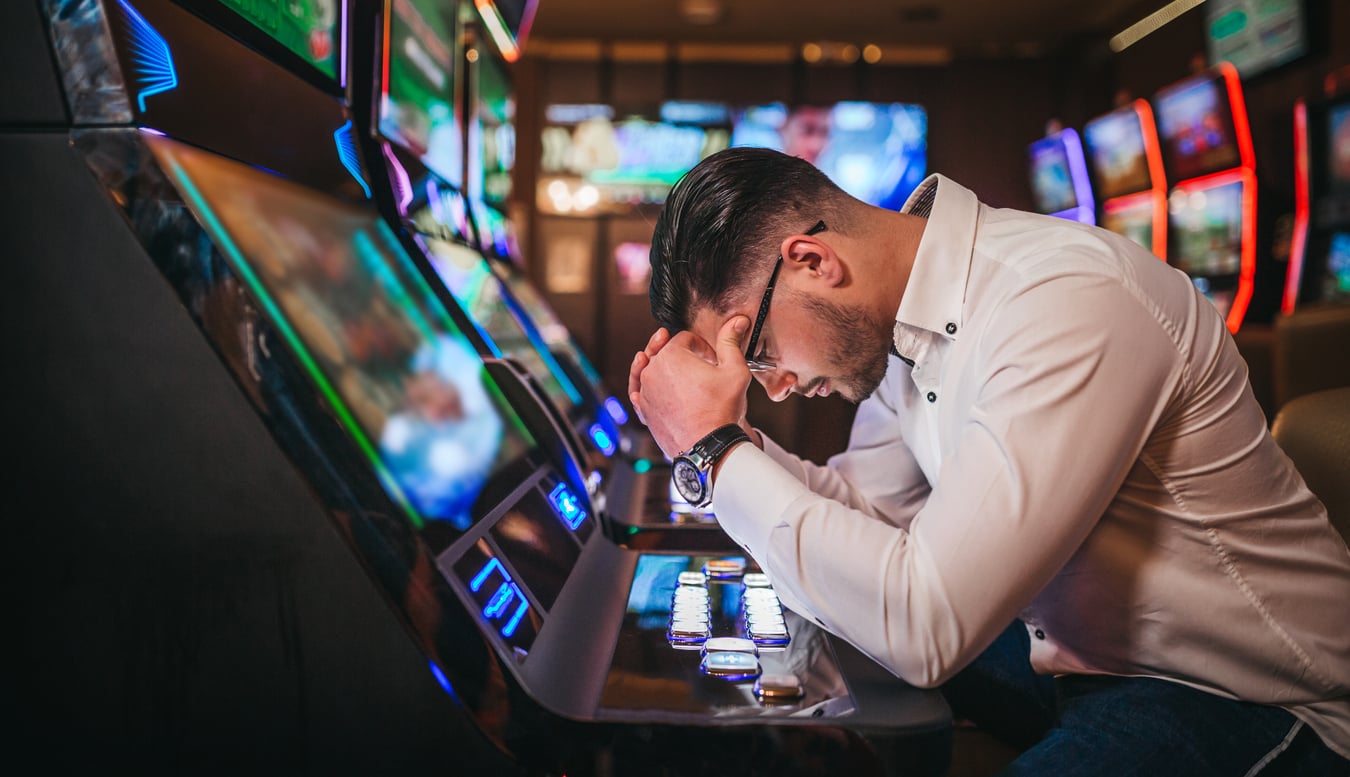 A man sits at a slot machine in a casino, leaning forward with his head in his hand, appearing stressed or upset—perhaps wishing he'd looked up some Vegas money-saving tips as colorful gaming machine lights glow around him.