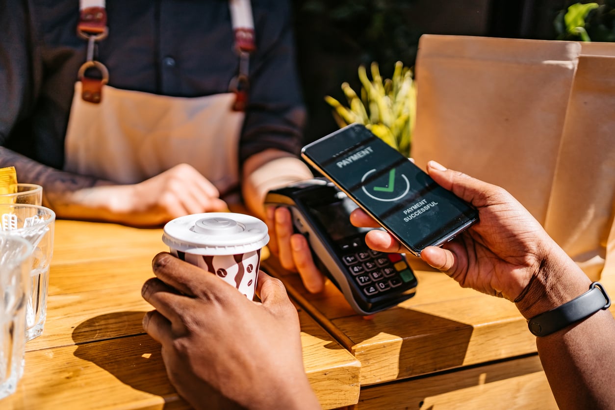 A person pays for a takeaway coffee using a smartphone at a counter, holding the coffee cup while another hand holds the phone near a card reader with a payment confirmation on the screen.