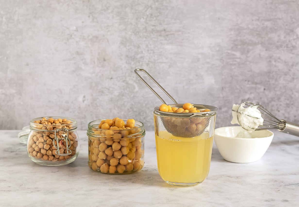 Three glass containers hold dried chickpeas, cooked chickpeas, and chickpea liquid (aquafaba), a popular egg substitute. A strainer holds chickpeas above the liquid, while a bowl with a whisk and whipped aquafaba sits in the background.