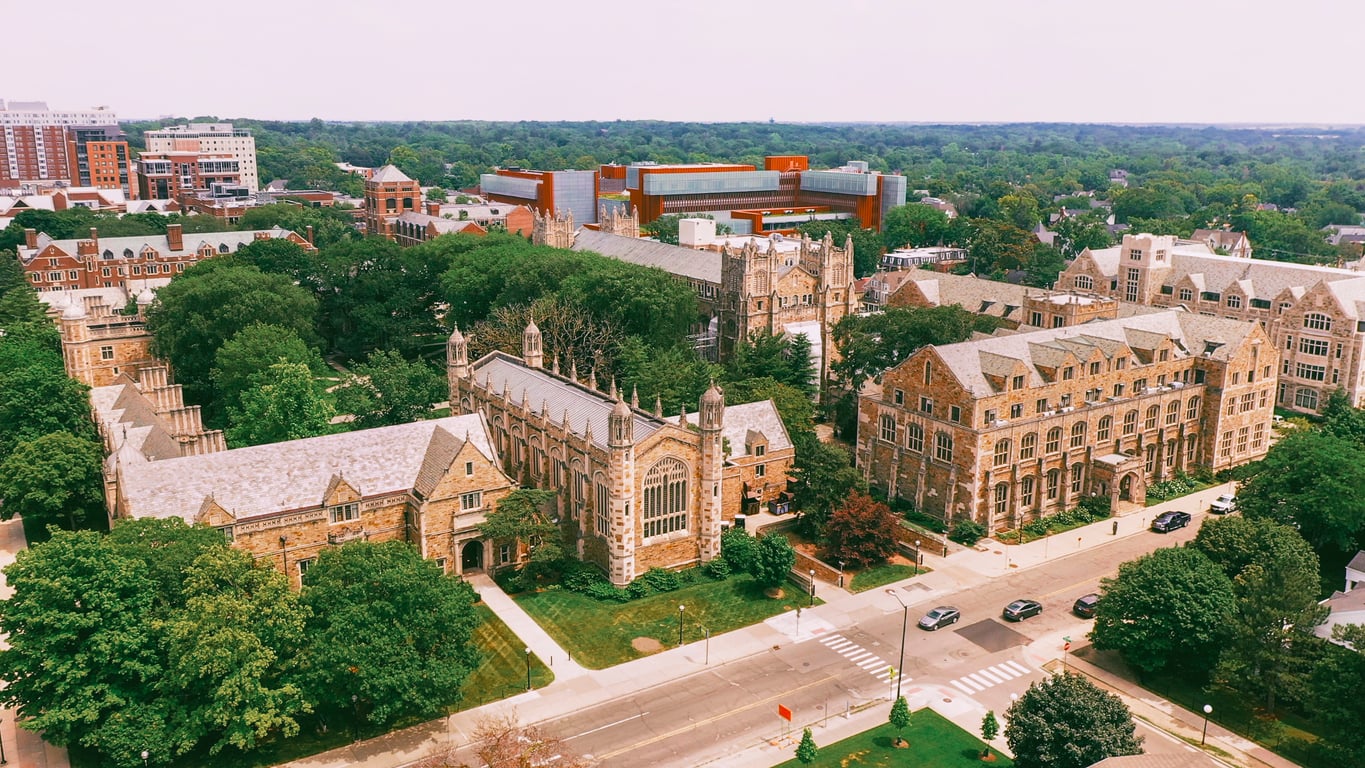 Aerial view of an affordable college town campus with historic gothic-style stone buildings, green trees, a modern building in the background, and a street with crosswalks and cars in the foreground.