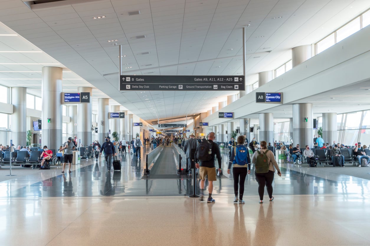 A spacious, convenient airport terminal with large windows, travelers walking and sitting, overhead signs showing gate directions, and a moving walkway in the center.
