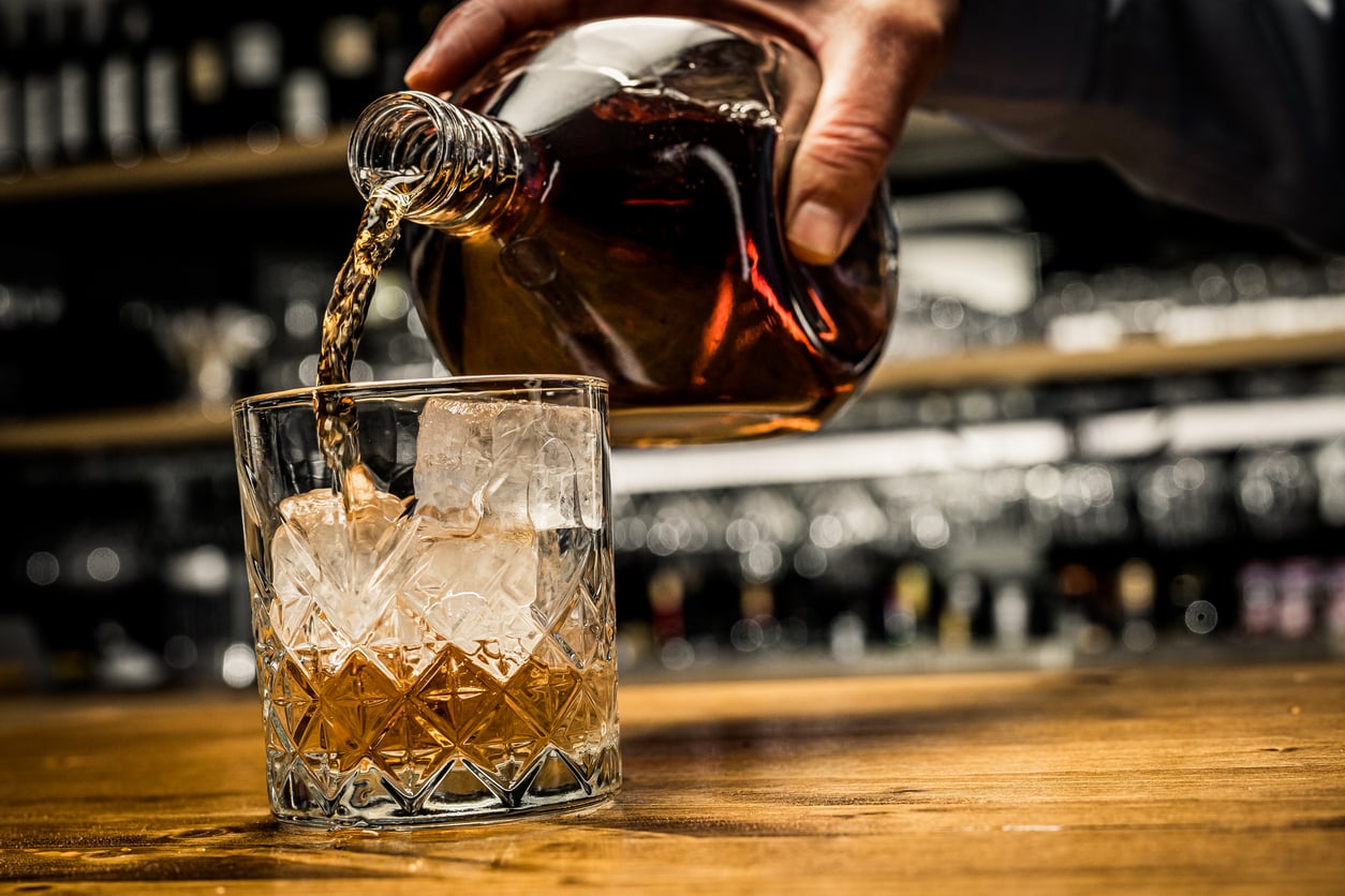 A hand pours amber-colored liquor from a glass bottle into a tumbler with ice cubes on a wooden bar, evoking the classic refinement of products men skip buying, with blurred bottles and glasses in the background.