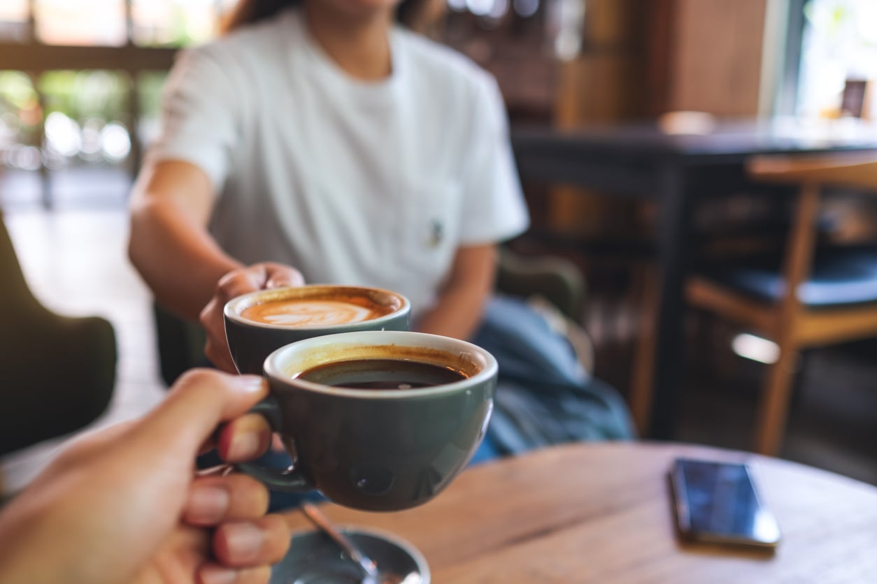 Two people clink coffee cups in a cozy café, savoring their favorite Coffee Brands. One cup holds black coffee, the other a latte. A blurred person in a white shirt sits in the background, with a table and a smartphone nearby.