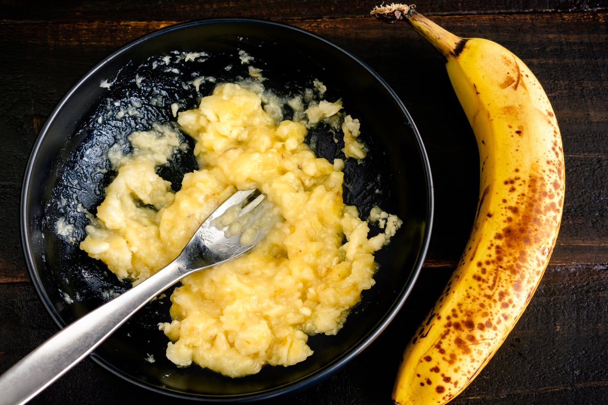 A black bowl with mashed banana and a fork inside, next to a ripe, spotted banana on a dark wooden surface—perfect for those seeking egg substitutes in baking recipes.