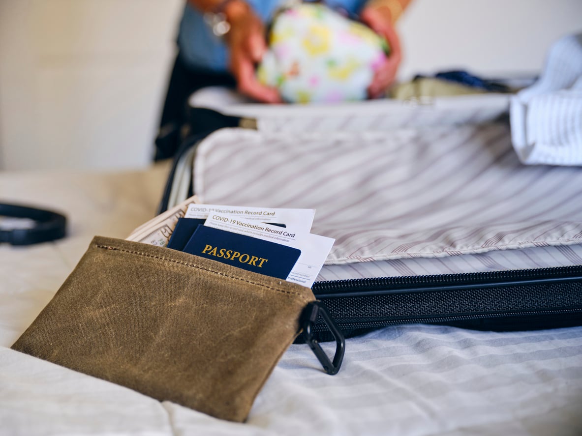 A close-up of a travel pouch with a passport and COVID-19 vaccination cards sticking out, placed on a bed next to an open suitcase being packed by a person in the background while staying at an Airbnb.