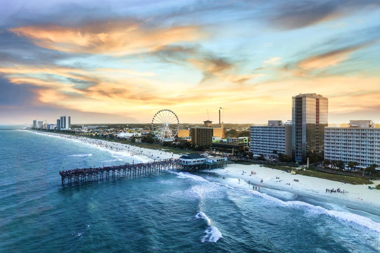 Aerial view of a beach city at sunset with a pier extending into the ocean, crowds on the sand, tall hotels lining the shore, and a large Ferris wheel near the coastline under a colorful sky.