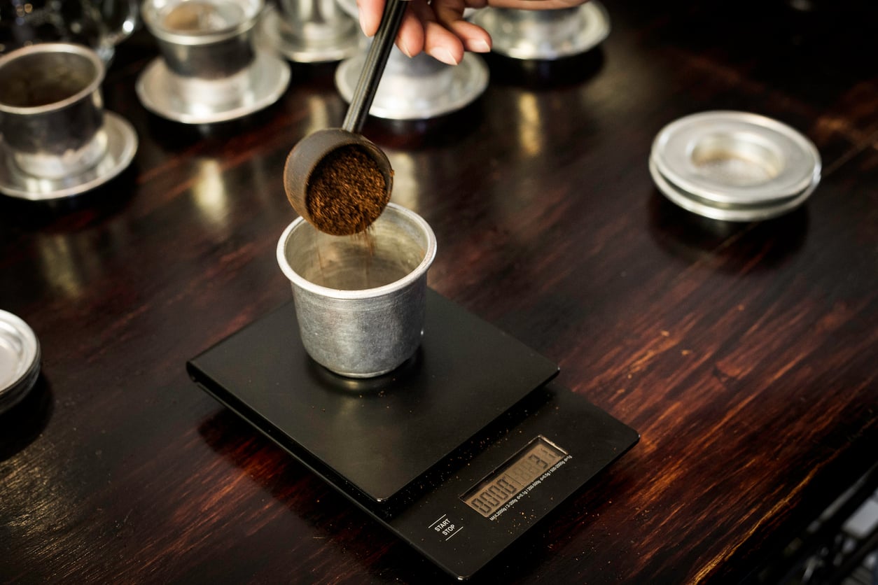 A hand scoops ground coffee into a metal filter placed on a digital scale atop a dark wooden surface, with several similar metal filters in the background.