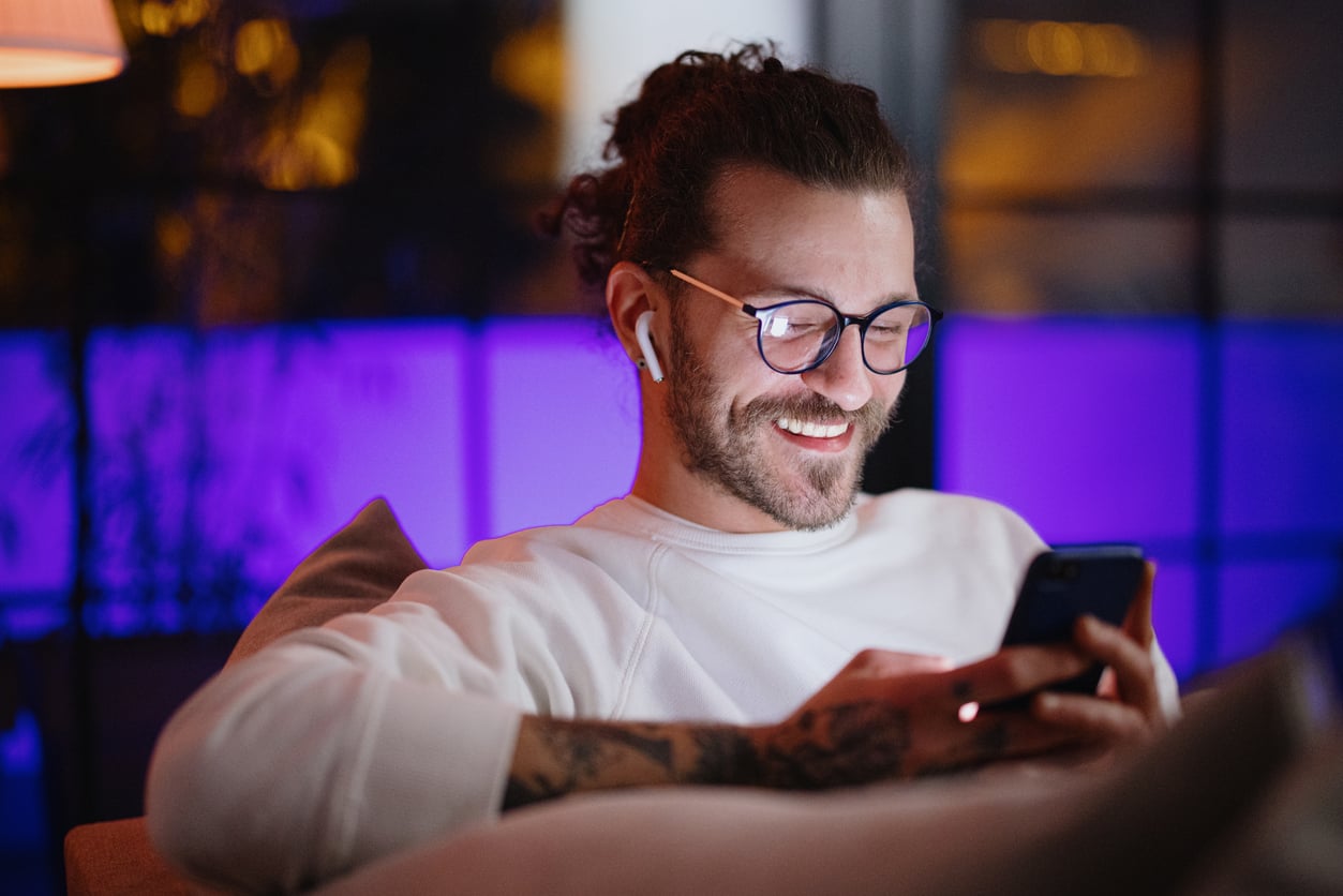 A man with glasses and wireless earbuds sits on a couch, smiling while looking at his smartphone—proof that some products men still buy never go out of style. He has a beard, long hair tied back, tattoos on his arm, and a softly lit purple background.