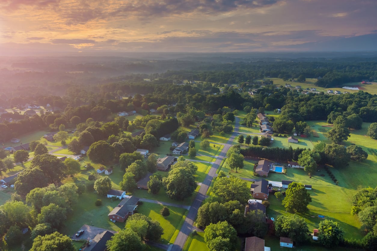 Aerial view of a suburban neighborhood at sunrise, with houses, green lawns, and trees lining both sides of a road. The landscape extends into a forested area under a colorful, partly cloudy sky.