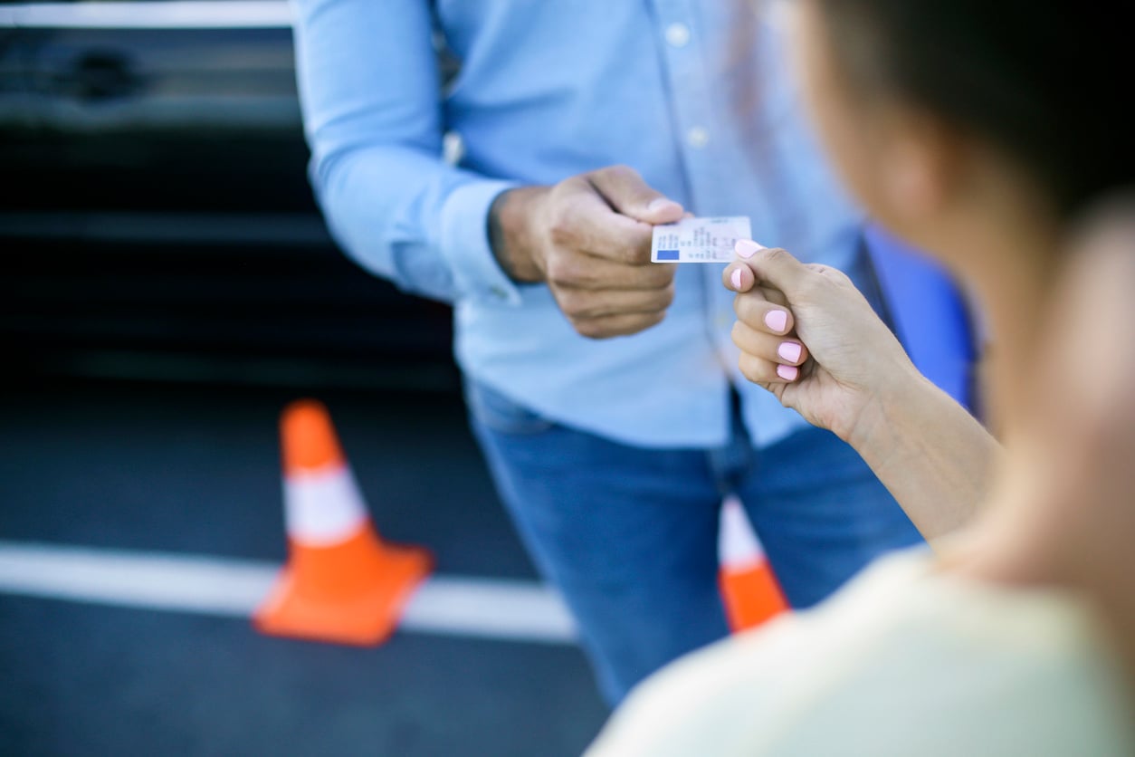 A person hands a small test device to another person outdoors, with orange traffic cones and a car visible in the background.