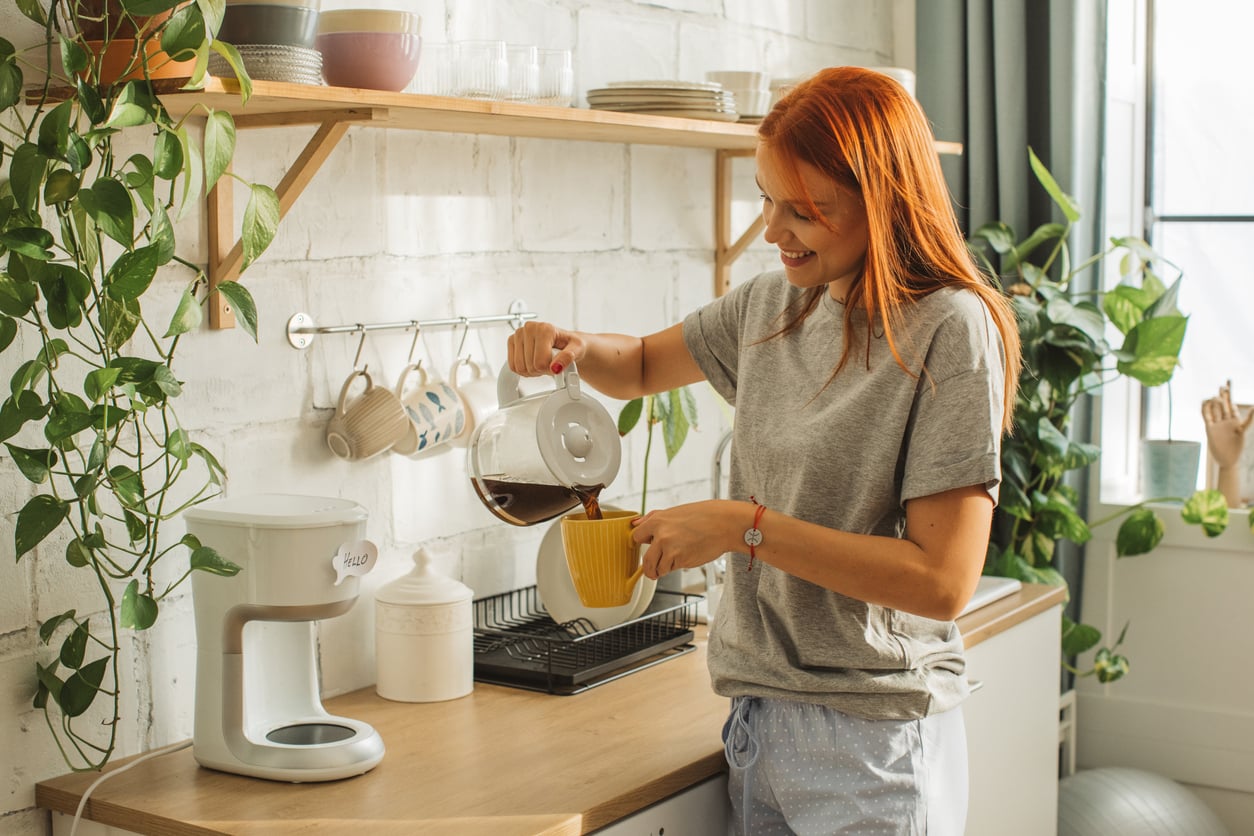 A woman with red hair smiles as she pours coffee from a glass carafe into a yellow mug in a bright, modern kitchen with plants and shelves holding dishes.