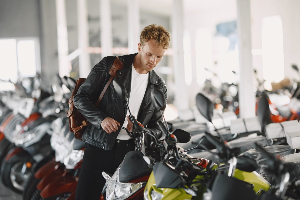 A young man with curly hair, wearing a black leather jacket and carrying a brown backpack, examines a green motorcycle—one of those products men skip buying—in a showroom filled with various motorcycles.